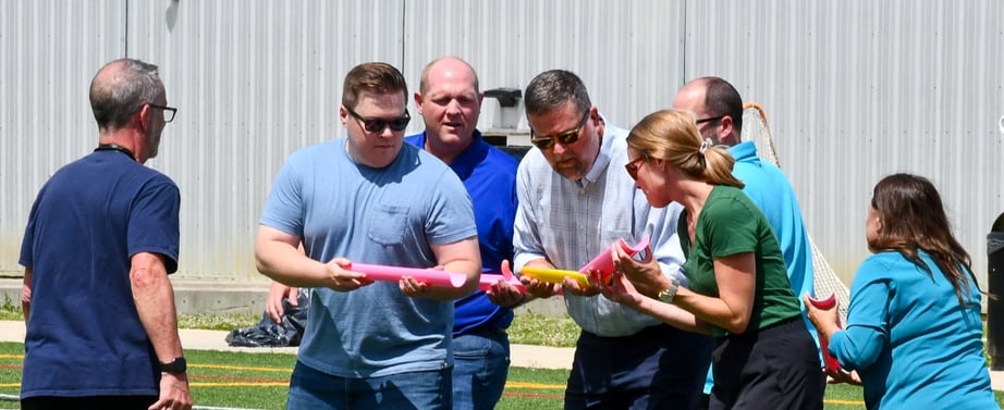A group of six people, holding colorful foam noodles, participates in an outdoor activity on a green athletic field, surrounded by a gray building and a soccer goal.