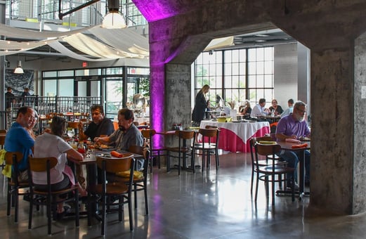 Pepple sit and eat inside a restaurant featuring concrete pillars, uplights, and upscale casual seating