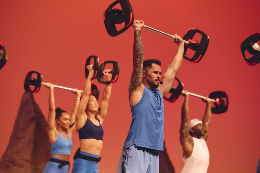 A group of fit people lift a barbell over their heads with straight arms.