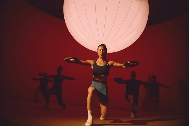 A group of fit people holding weight plates in both hands lift their arms to a 90 degree angle while in a deep lunge.