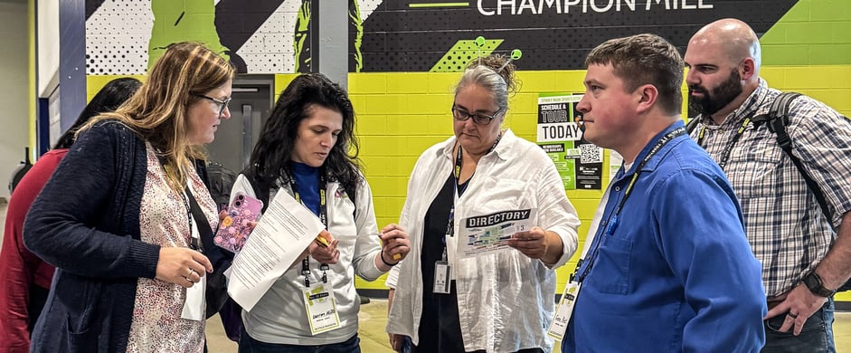 A group of 6 adults stand in a group, looking at papers with information. They are pensive in thought.
