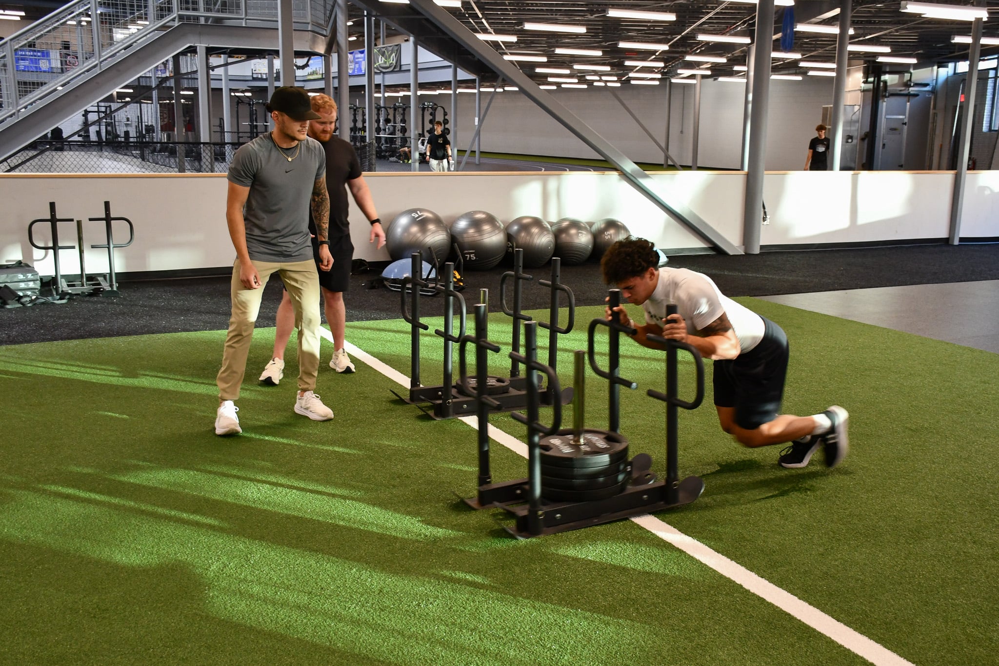 An athlete is pushing a fitness sled with weights on it. Two coaches stand to the side watching the athlete.