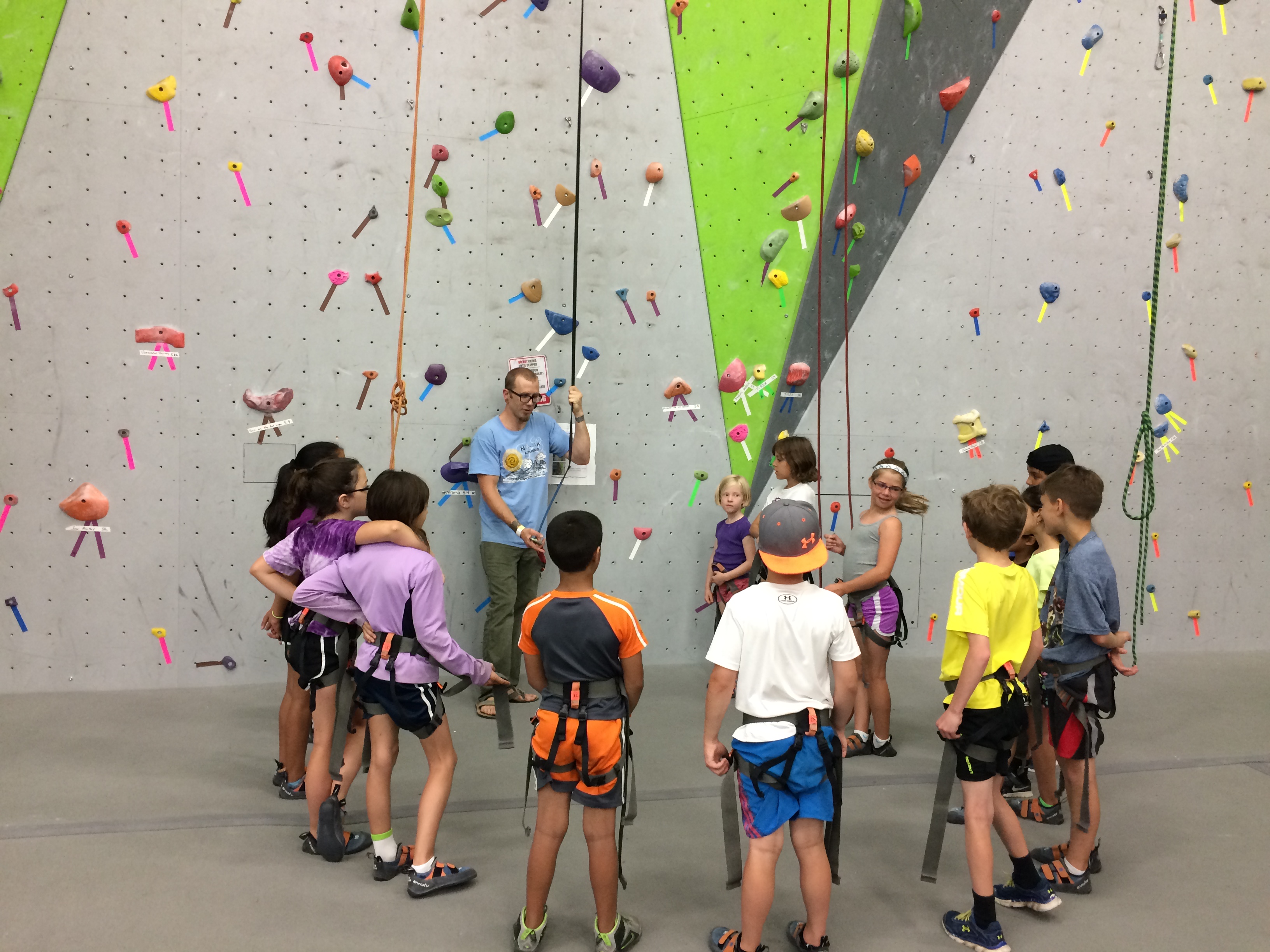 Children standing in a semi-circle around a rock wall and a rock wall climbing instructor at a summer camp in Hamilton, Ohio
